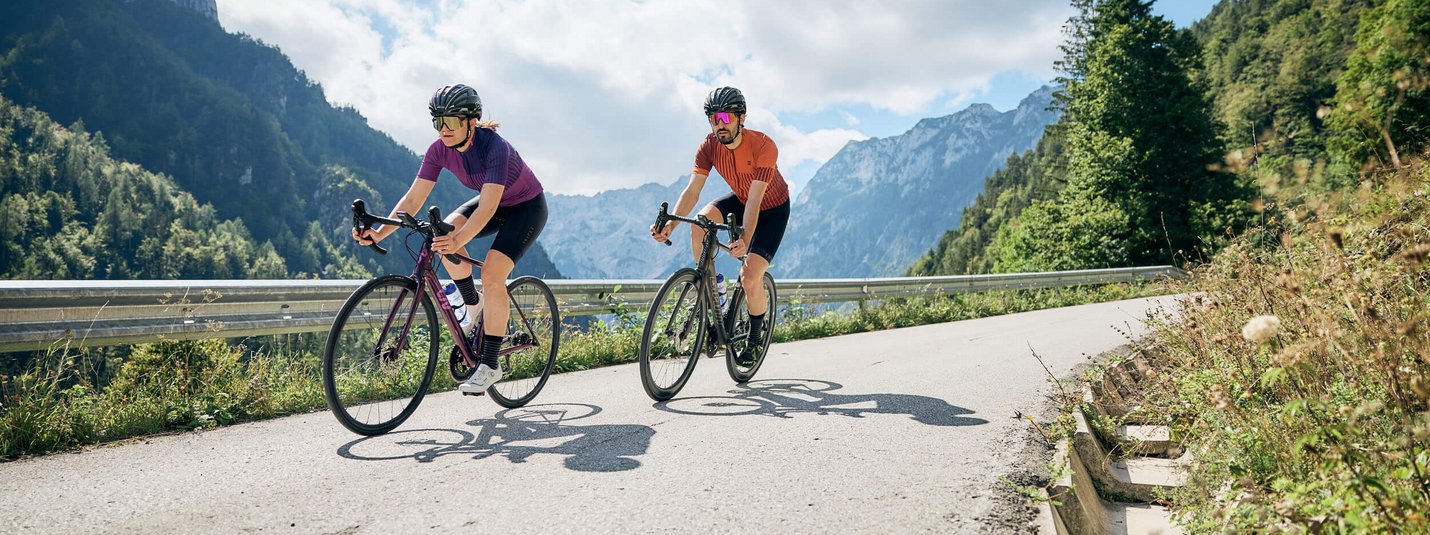 Two cyclists riding on mountain road under sunny sky with clouds