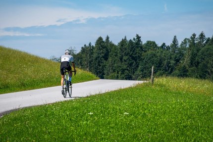 Ciclista su strada di campagna circondata da prati verdi e alberi con cielo azzurro