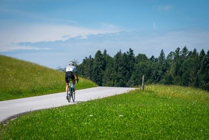 Cyclist on country road surrounded by green fields and trees under blue sky