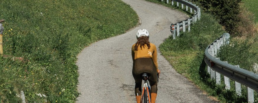 Fahrradfahrerin auf kurviger Straße durch grüne Hügel unter bewölktem Himmel