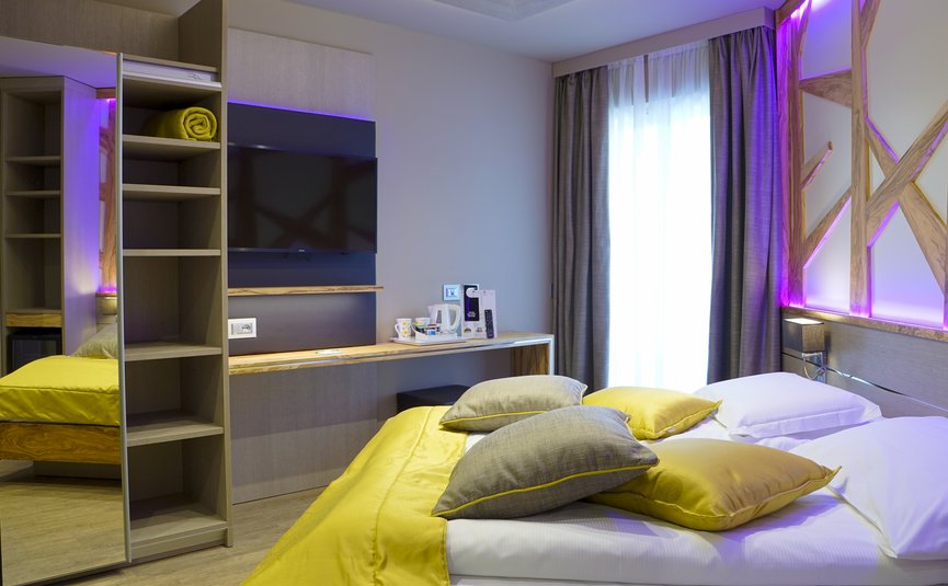 Modernes Hotelzimmer mit gelber Bettdecke und Kissen, TV und Schrank mit Spiegel