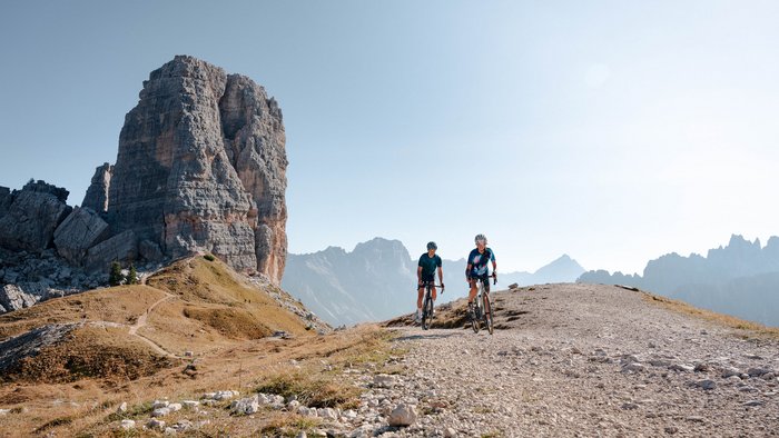 Two cyclists on mountain trail with large rock formation and mountain backdrop