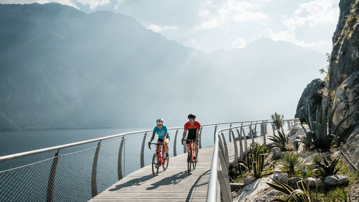 Zwei Radfahrer auf einem Steg am See mit Bergen im Hintergrund