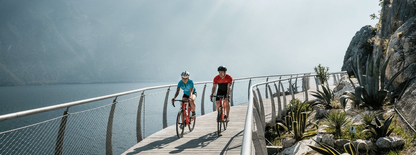 Zwei Radfahrer auf einem Steg am See mit Bergen im Hintergrund