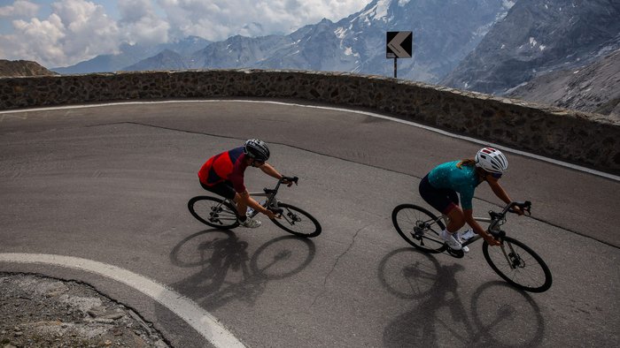 Two cyclists riding a tight mountain road with snow-capped peaks in the background