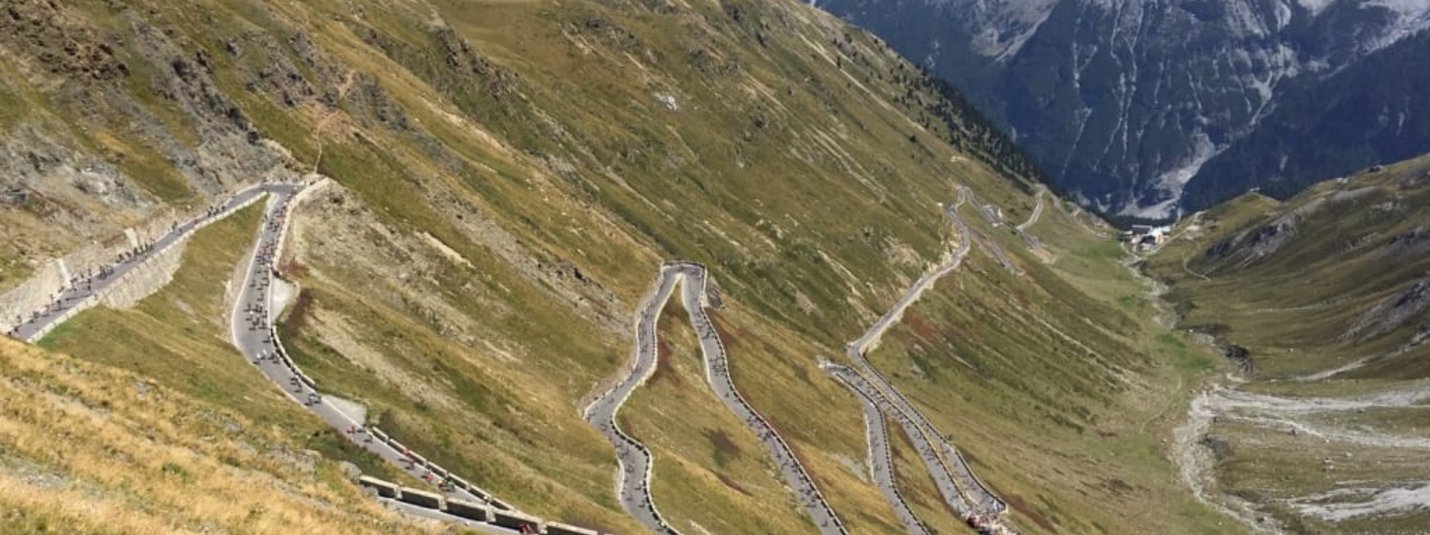 Strada montana a tornanti con ciclisti e cime innevate sullo sfondo