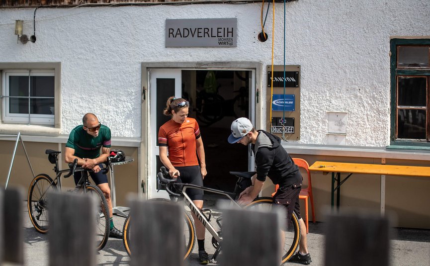 Two cyclists and a mechanic at bike rental outside building with signs