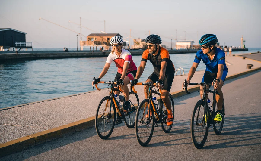 Vacanze in bici da corsa sulla costa adriatica © Alex Moling Tre ciclisti con abbigliamento colorato pedalano lungo il lungomare al chiaro cielo