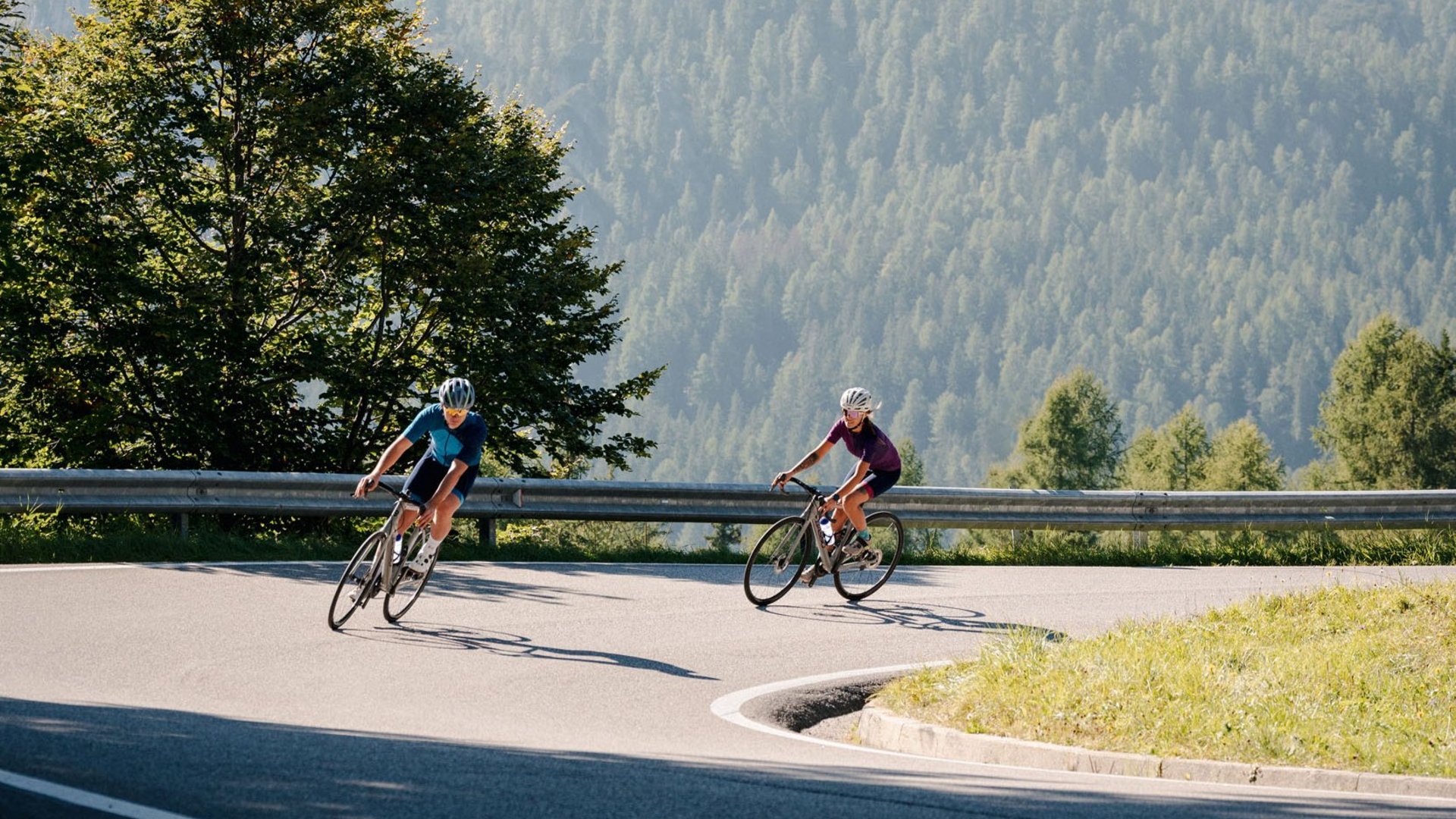 Zwei Radfahrer fahren auf kurviger Bergstraße mit bewaldeten Bergen im Hintergrund