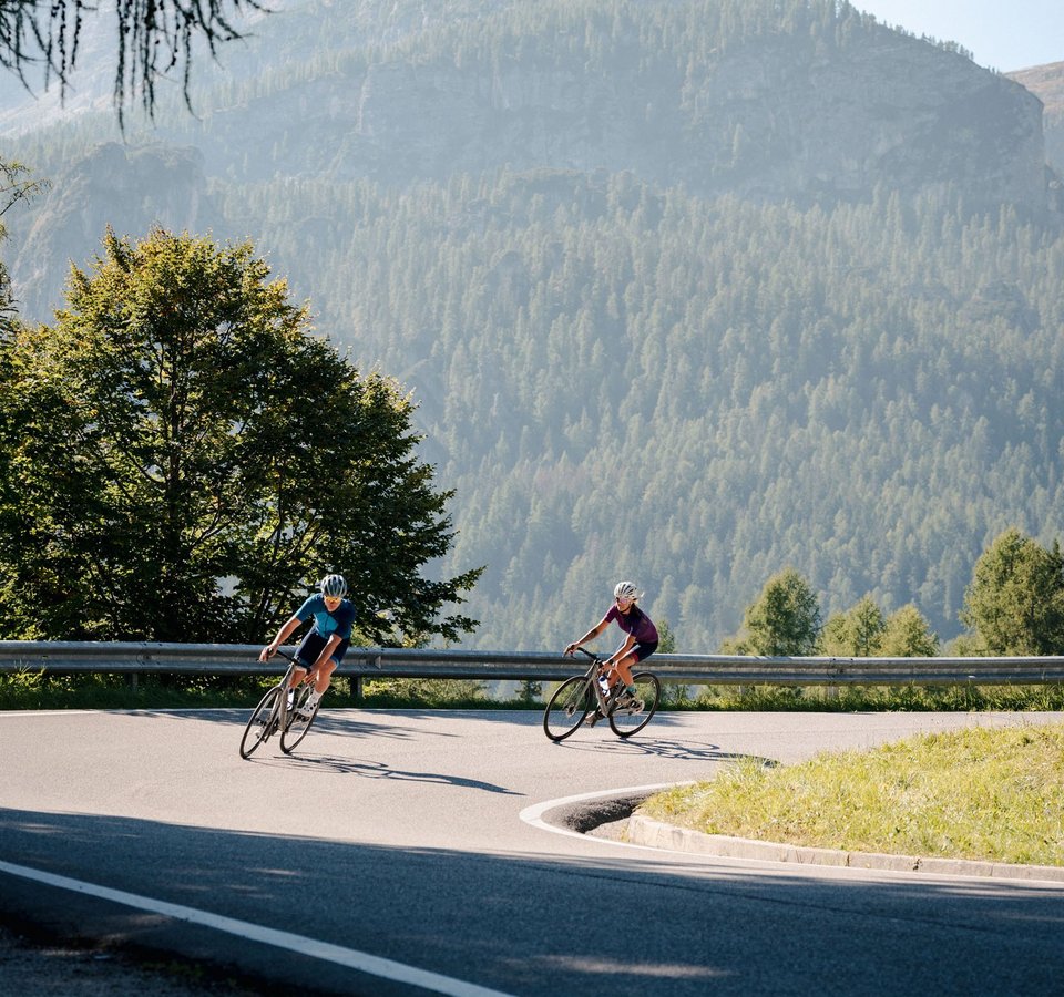 Zwei Radfahrer fahren auf kurviger Bergstraße mit bewaldeten Bergen im Hintergrund