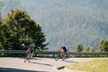 Zwei Radfahrer fahren auf kurviger Bergstraße mit bewaldeten Bergen im Hintergrund