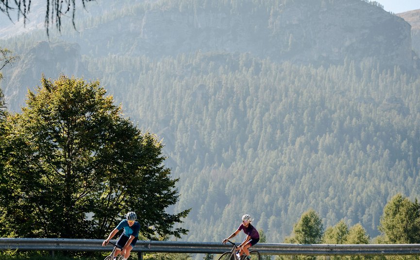 Two cyclists riding on a winding mountain road with forested mountains behind