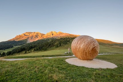 Reiter Joch © Günther Pichler - EyetotheDolomites Obereggen Holzskulptur in Form eines Eies auf einer Bergwiese bei Sonnenuntergang