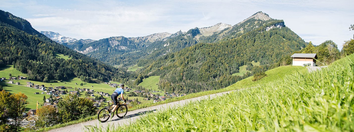 Radfahrer auf Bergstraße mit Dorf und Bergen im Hintergrund