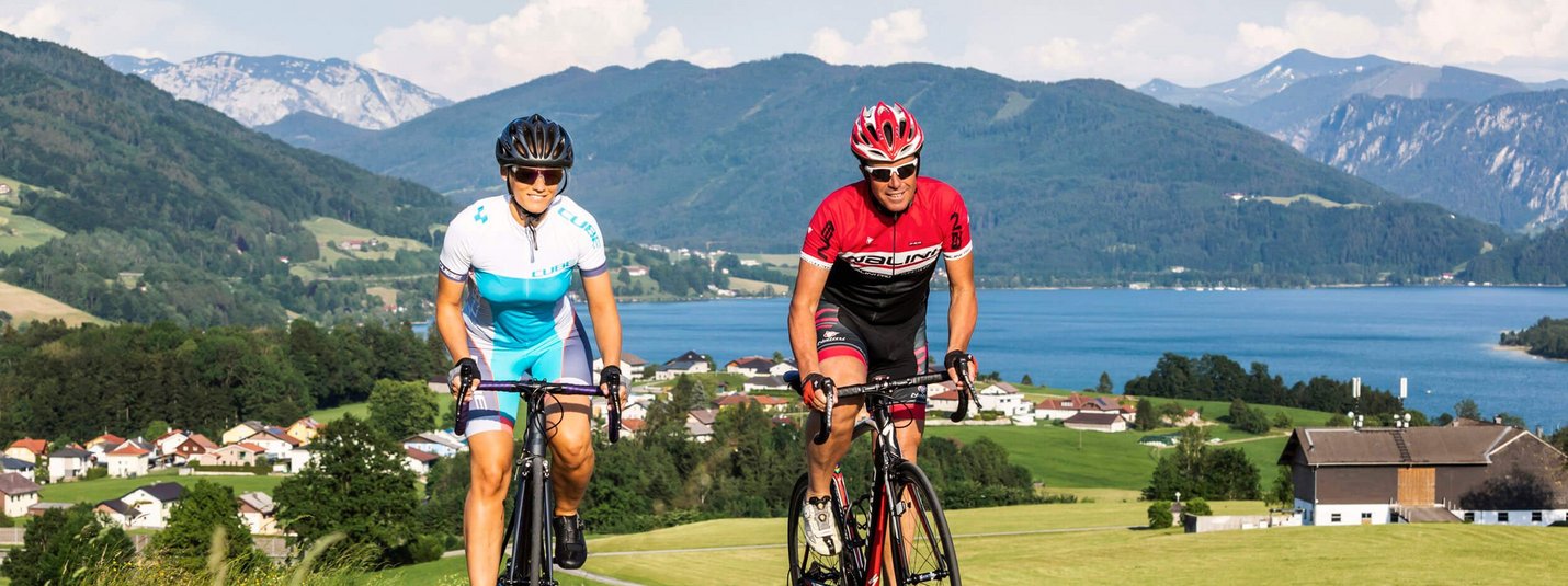Two cyclists on a country road with mountains and lake in background