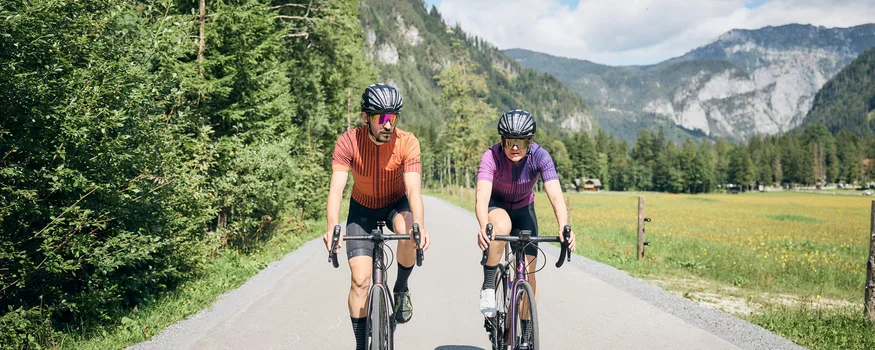 Zwei Radfahrer auf Landstraße vor bewaldeten Bergen bei sonnigem Wetter
