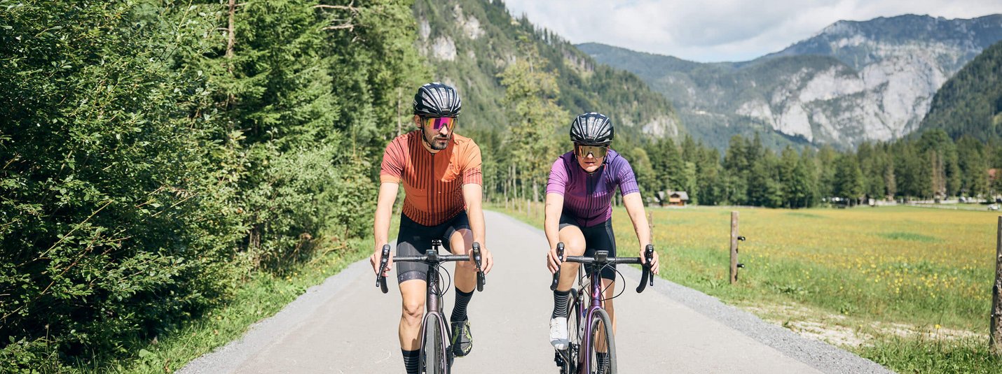 Due ciclisti su strada di campagna davanti a montagne boschive con cielo soleggiato