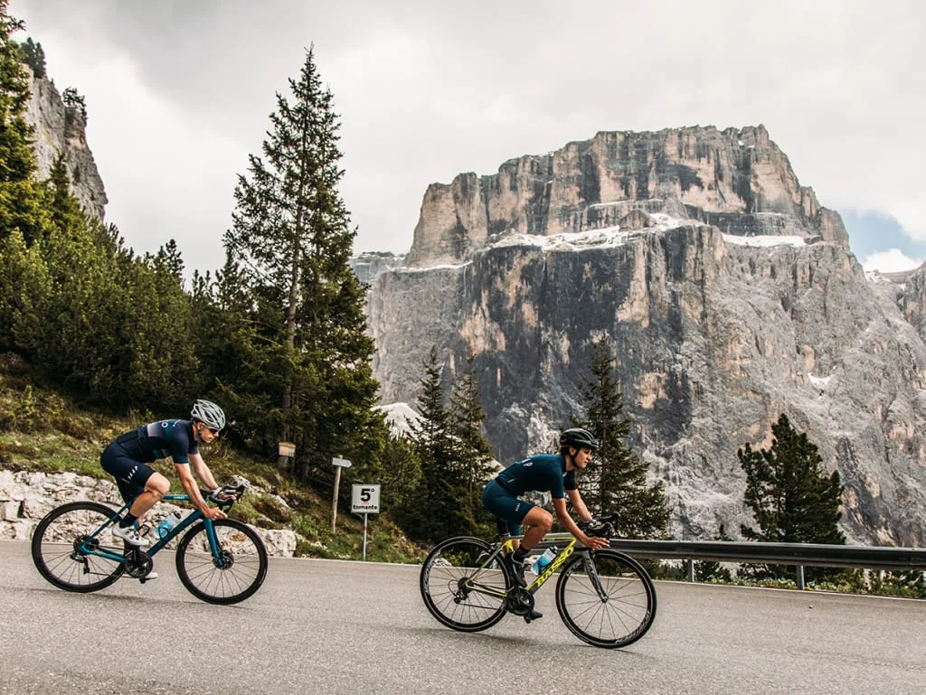 Two cyclists riding uphill on a road with mountains in the background