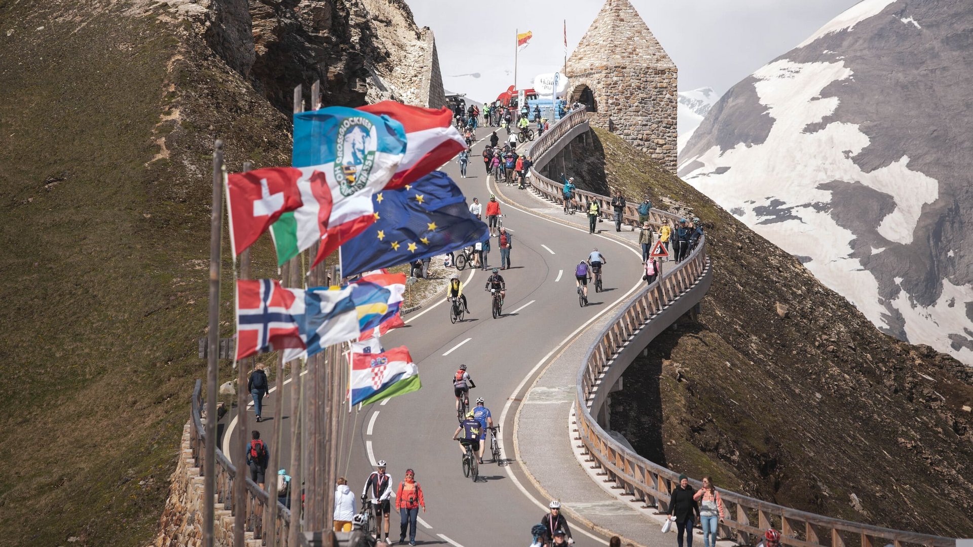 29. Glocknerkönig am 7. Juni 2026 © Kovac György Tourismusverband BRUCK FUSCH GROSSGLOCKNER Fahrradfahrer und Fußgänger auf einer Bergstraße mit Flaggen und Schneegipfeln