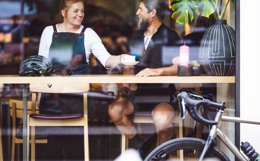 Man and woman laughing and drinking coffee in café, bicycle outside window