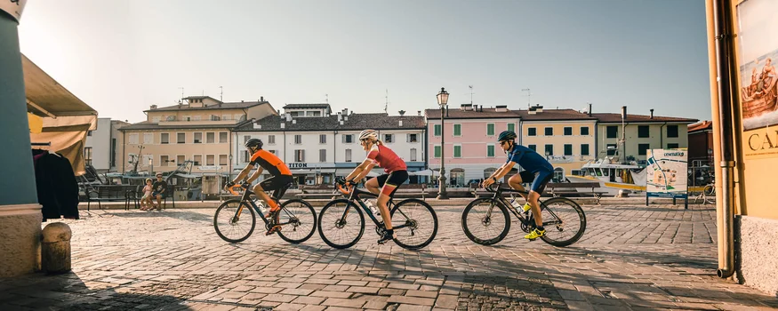 Tre ciclisti pedalano in una piazza soleggiata di una piccola città