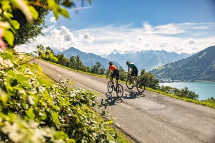 Zwei Radfahrer fahren auf Bergstraße mit Blick auf See und Berge bei sonnigem Wetter