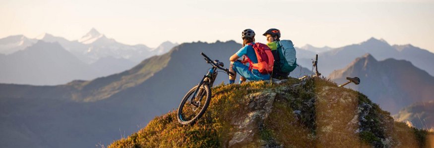 Zwei Radfahrer mit Rucksäcken sitzen auf einem Berggipfel mit Blick auf Berge