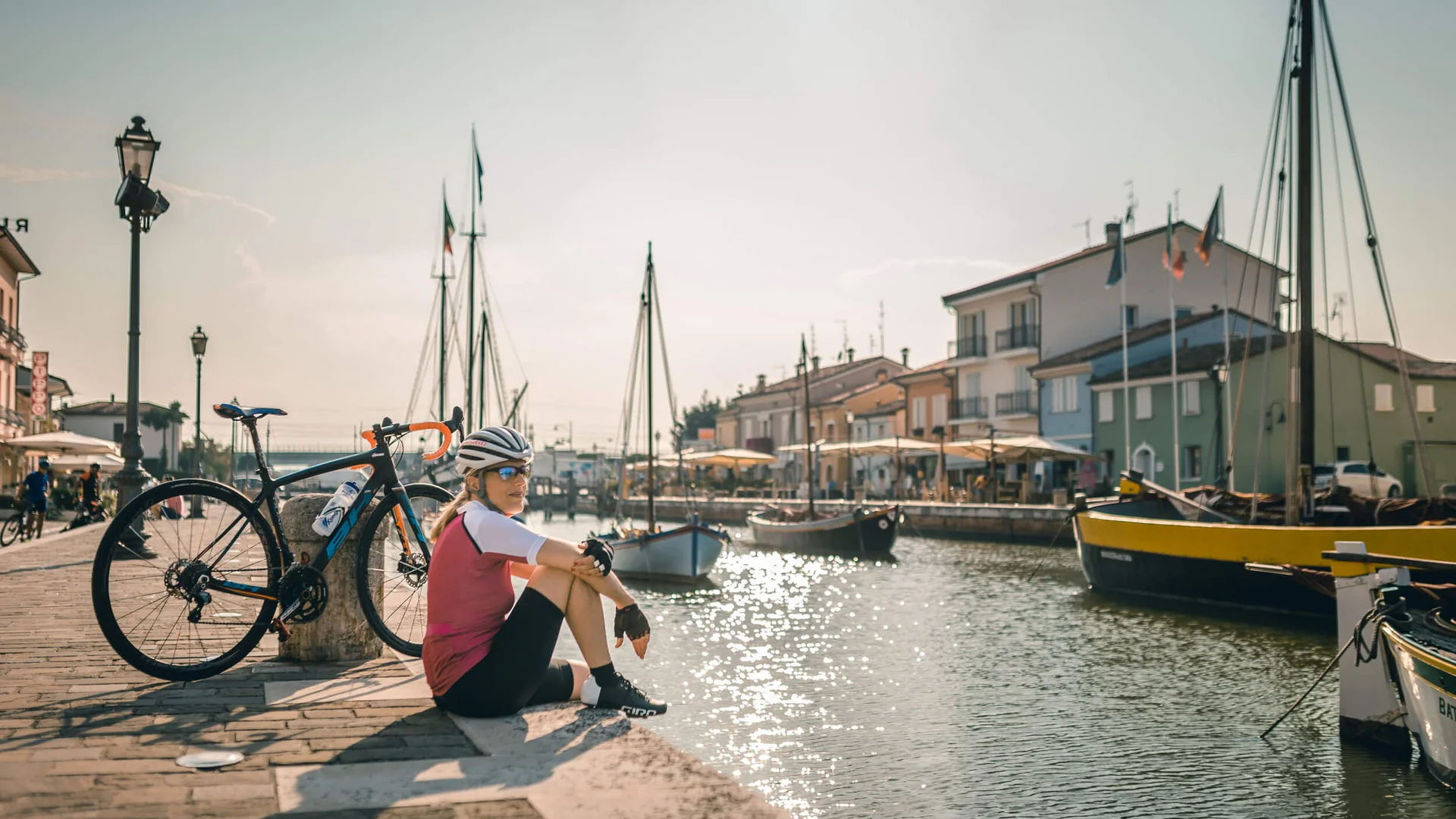 Adriatische Küste: Rennradurlaub mit Dolce Vita © Alex Moling Radfahrerin sitzt am Hafen mit Booten und Fahrrad im Sonnenschein