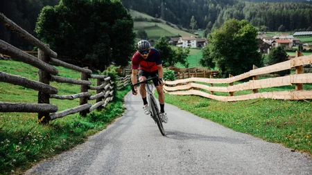 Mann fährt Rennrad auf Landstraße mit Holzzäunen und Bergen im Hintergrund
