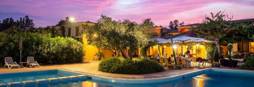 Evening by lit pool with terrace and people dining under pink purple sky