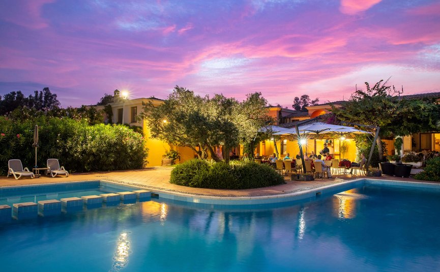 Evening by lit pool with terrace and people dining under pink purple sky