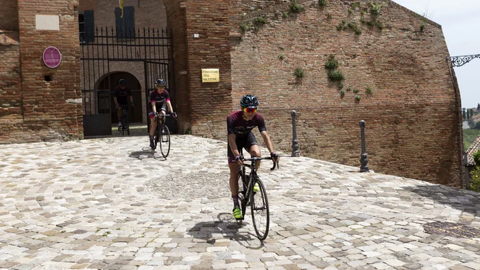 Adriatische Küste: Rennradurlaub mit Dolce Vita © Riccardo Rocchi Radfahrer fahren durch ein altes Tor in einer gepflasterten Straße