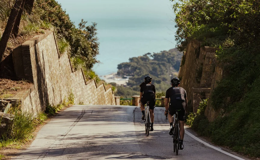 Zwei Radfahrer fahren auf einer Straße zum Meer mit bewaldeten Hügeln