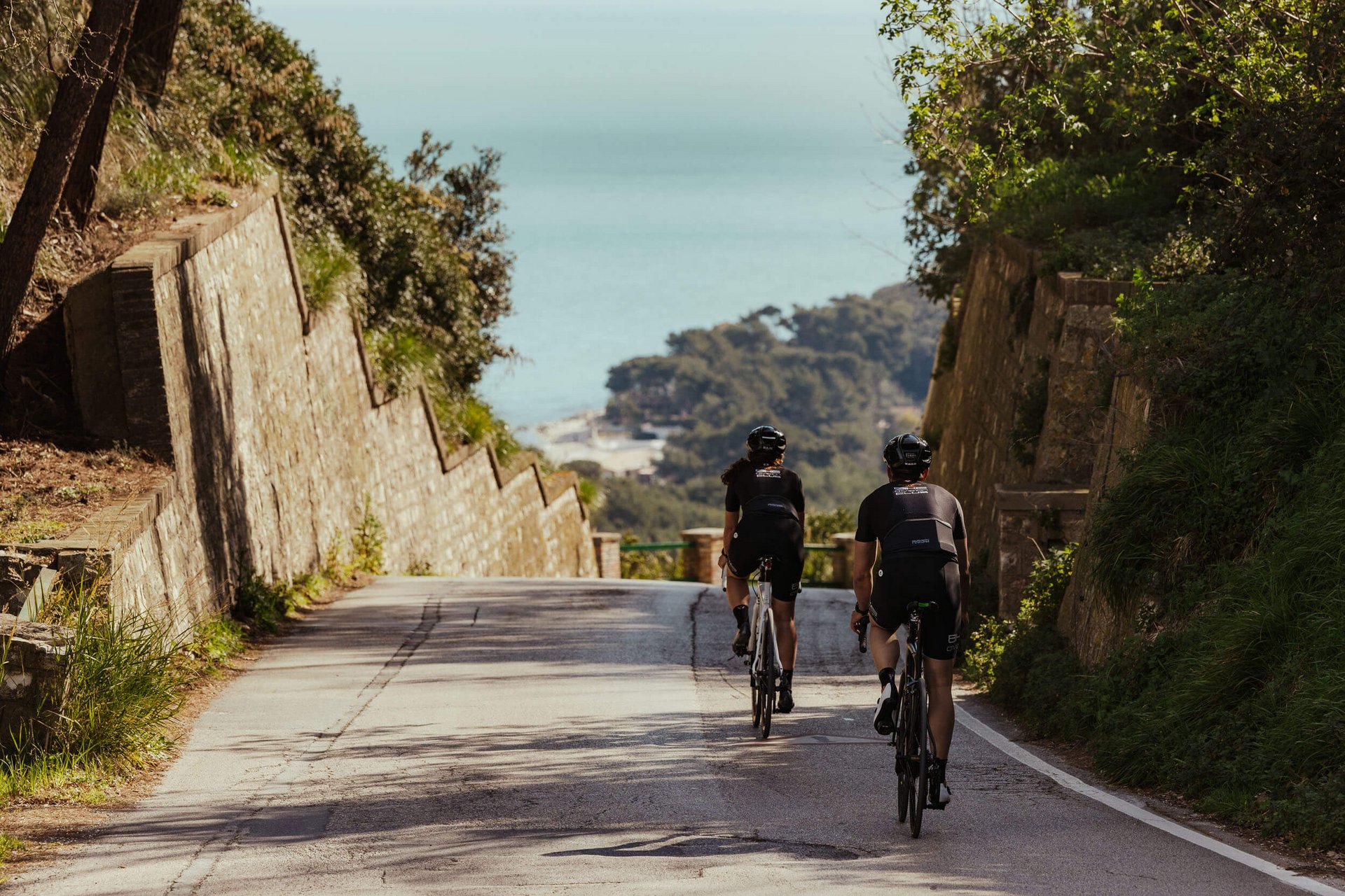 Roadbike Holidays © Alex Moling Zwei Radfahrer fahren auf einer Straße zum Meer mit bewaldeten Hügeln