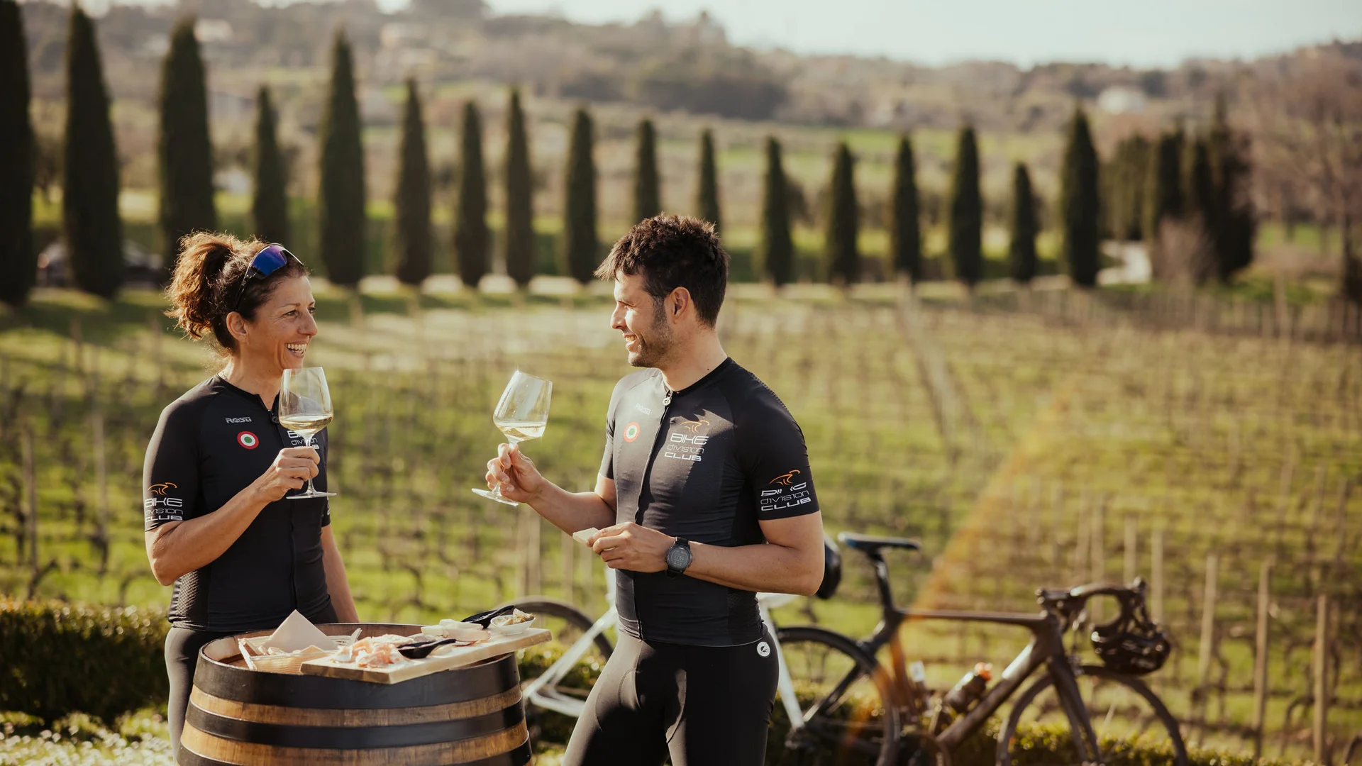 Two cyclists enjoying wine and snacks at a barrel in a vineyard
