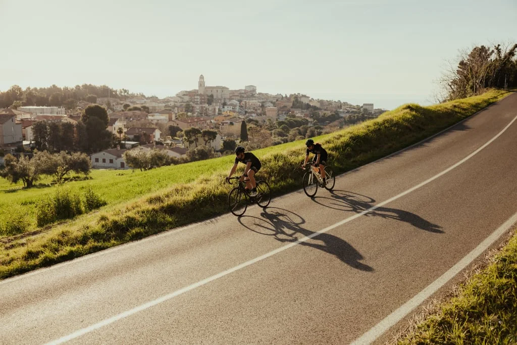 Exploring Marche Region by Bike Two cyclists ride along a road with a town in the background