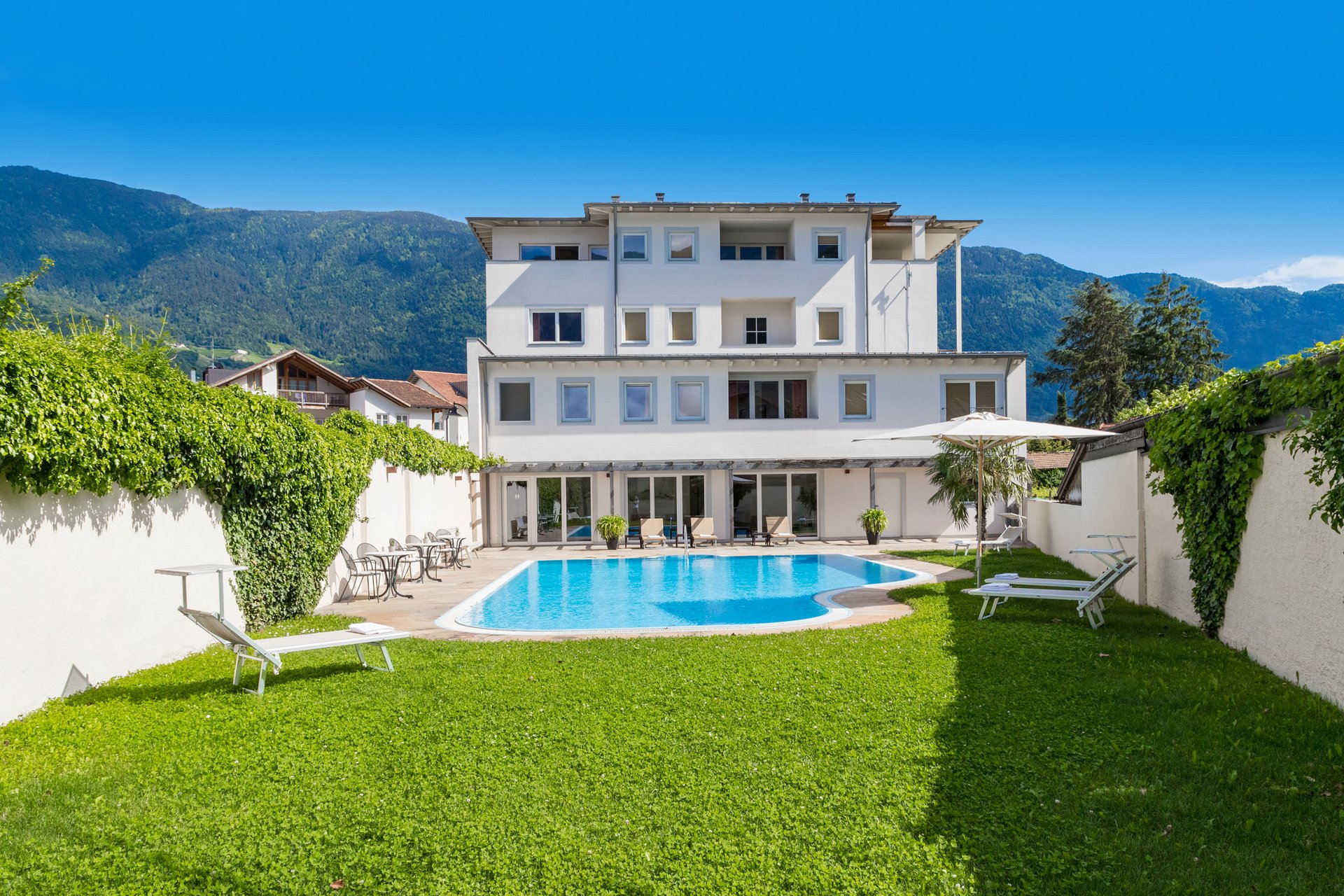 White hotel with outdoor pool, lounge chairs, and mountain view under clear sky