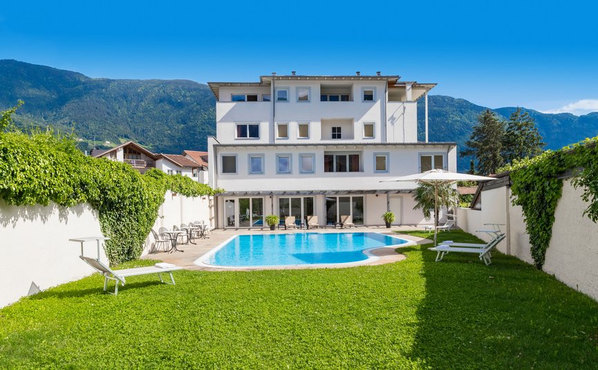 Weißes Hotel mit Außenpool, Liegestühlen und Bergblick bei klarem Himmel