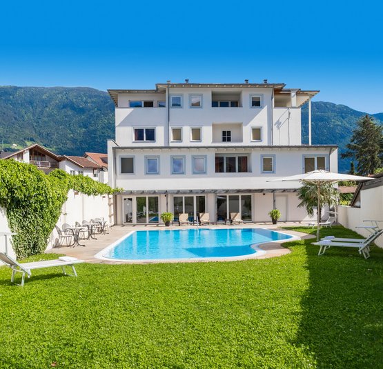 Hotel bianco con piscina esterna, lettini e vista sulle montagne sotto un cielo sereno