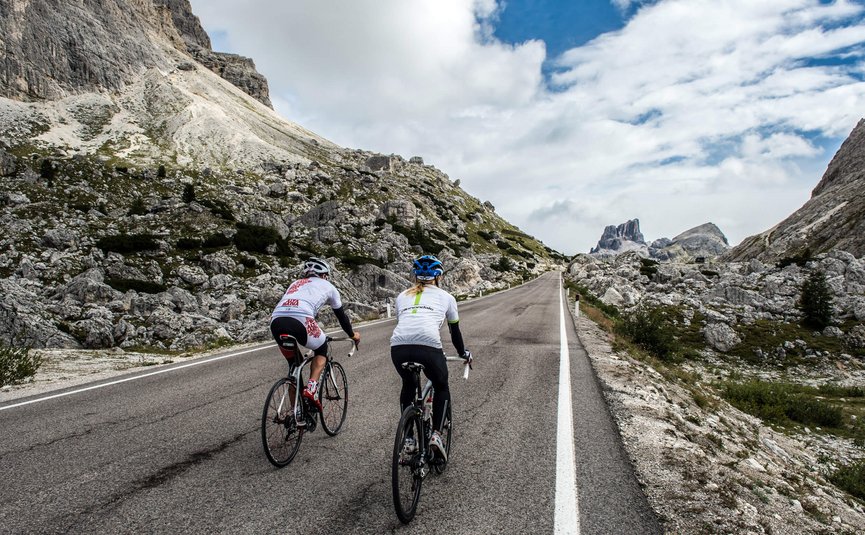 Two cyclists riding uphill on mountain road with rocky landscape and cloudy sky.