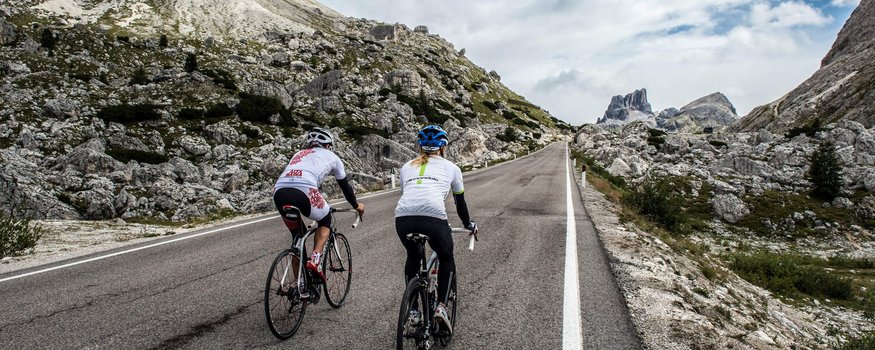 Twee fietsers rijden omhoog op bergweg met rotsachtig landschap en bewolkte lucht.