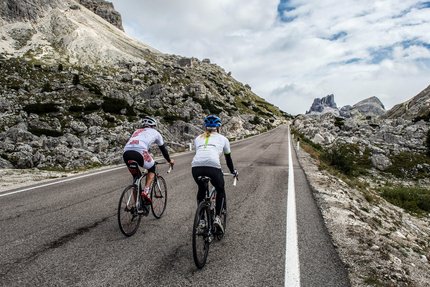 Two cyclists riding uphill on mountain road with rocky landscape and cloudy sky.