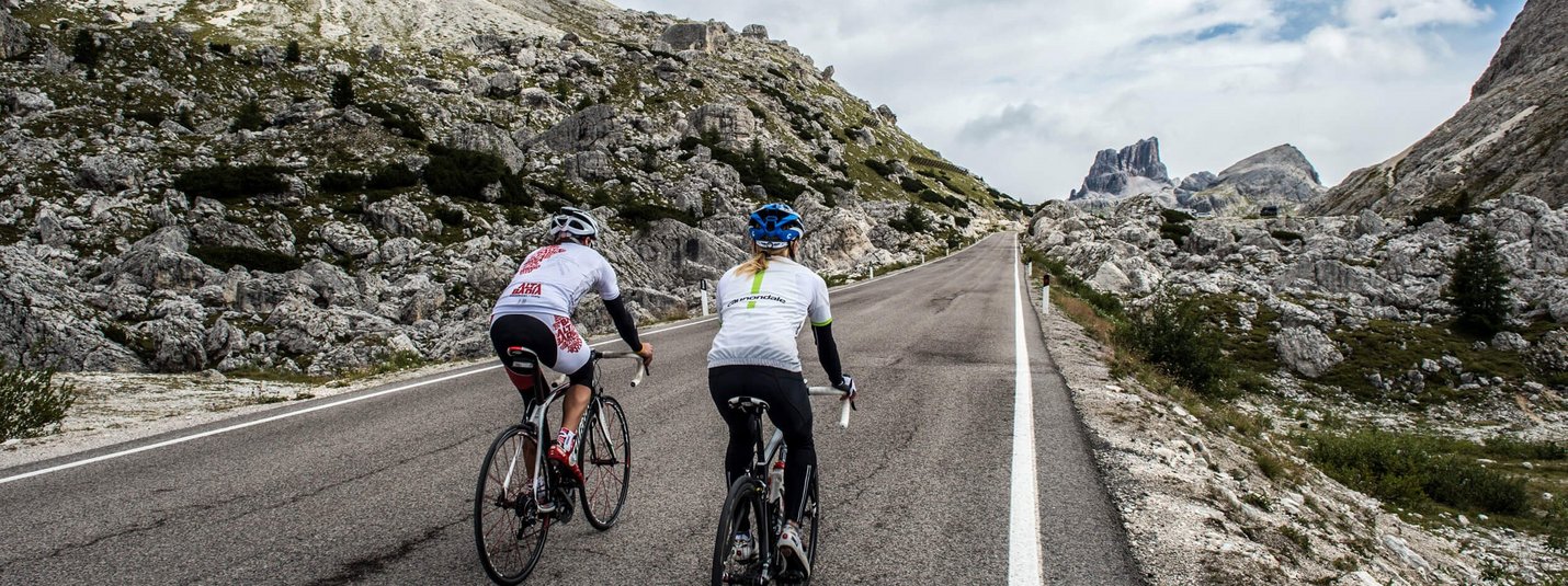 Due ciclisti pedalano su una strada di montagna con rocce e cielo nuvoloso.