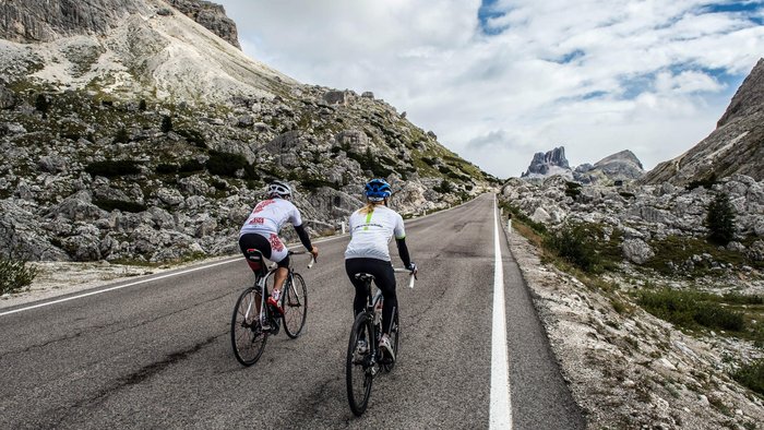 Two cyclists riding uphill on mountain road with rocky landscape and cloudy sky.