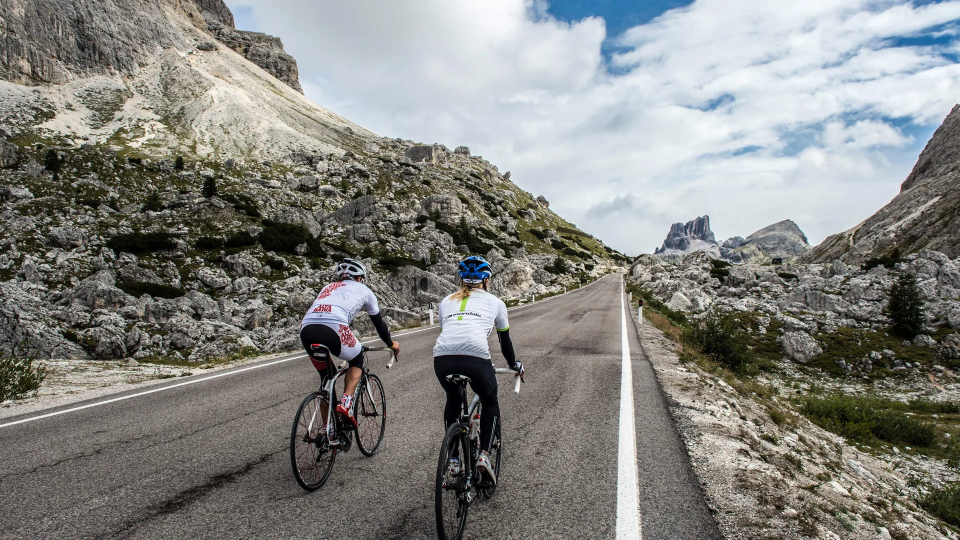 Due ciclisti pedalano su una strada di montagna con rocce e cielo nuvoloso.