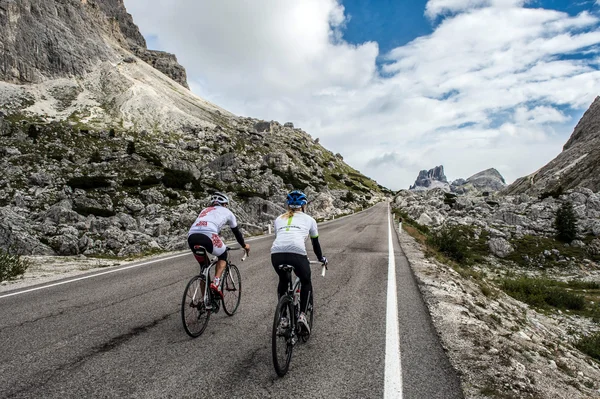 Italy © Paola Finali Two cyclists riding uphill on mountain road with rocky landscape and cloudy sky.