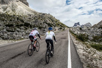 Two cyclists riding uphill on mountain road with rocky landscape and cloudy sky.