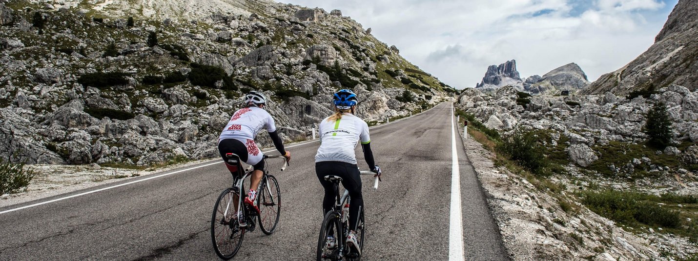 Two cyclists riding uphill on mountain road with rocky landscape and cloudy sky.