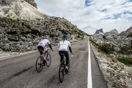 Zwei Radfahrer fahren eine Bergstraße mit Felsen und wolkigem Himmel.