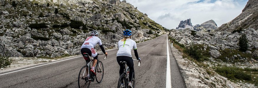 Zwei Radfahrer fahren eine Bergstraße mit Felsen und wolkigem Himmel.